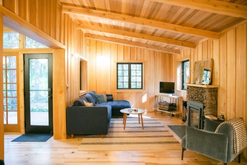 Cozy wooden living room with a sofa, coffee table, TV, and stone fireplace, surrounded by large windows.