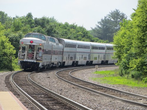 A vintage train curves around a bend, surrounded by lush greenery and a clear sky.
