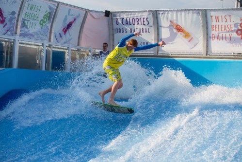 A surfer in a yellow wetsuit rides a wave on a flowboard at a water park.