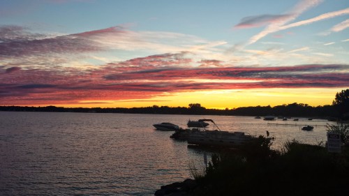 A serene lake at sunset, with colorful clouds reflecting on the water and boats moored along the shore.