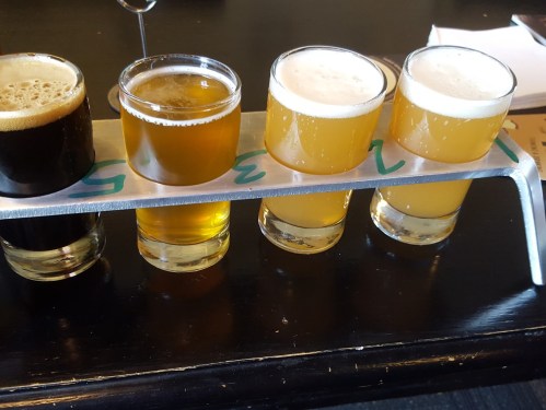 A flight of four beer samples on a metal holder, featuring dark and light brews in small glasses.