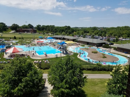 Aerial view of a vibrant outdoor pool area with slides, sunbathers, and green lawns under a clear blue sky.