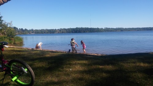 Children playing by a calm lake on a sunny day, with trees in the background and a bike in the foreground.