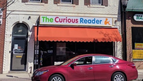 A storefront with a colorful sign reading "The Curious Reader" and a red car parked in front.