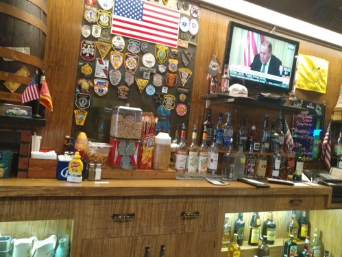 A bar counter with various liquor bottles, condiments, and a TV displaying news, surrounded by badges and an American flag.