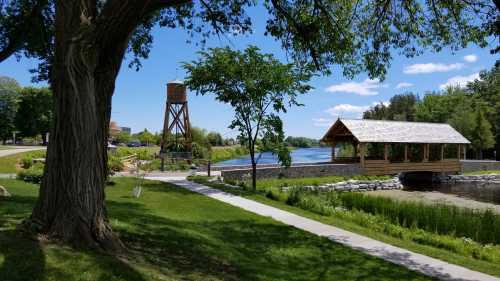 A scenic park with a wooden bridge, water tower, and a river, surrounded by lush greenery and blue skies.