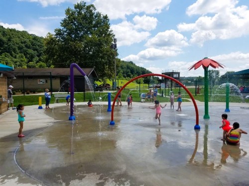 Children play in a splash pad with colorful water features under a sunny sky and fluffy clouds.
