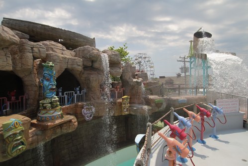 A water park scene featuring colorful tiki statues, waterfalls, and water spray features against a cloudy sky.