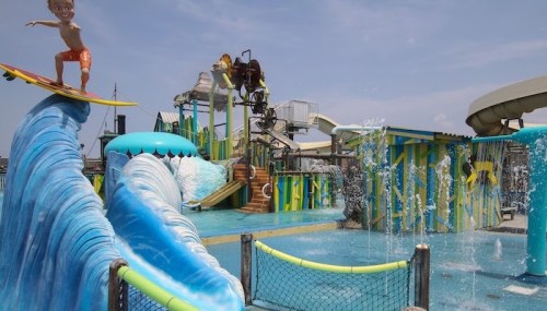A vibrant water park scene featuring a child surfing on a wave with colorful slides and water features in the background.