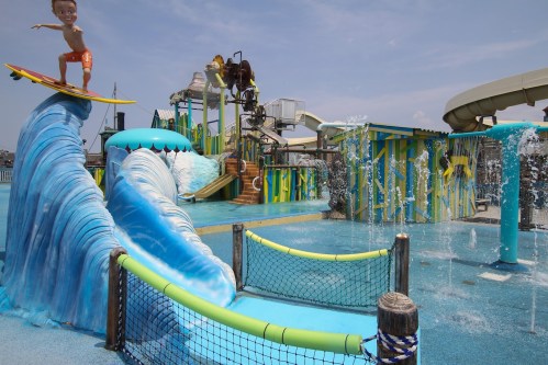 A colorful water park featuring slides, fountains, and a surfboarder statue riding a wave.