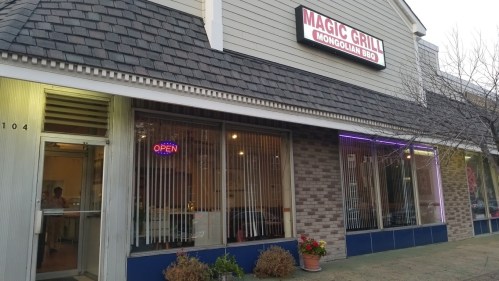 Exterior of Magic Grill, a Mongolian BBQ restaurant, featuring an "Open" sign and colorful lights.