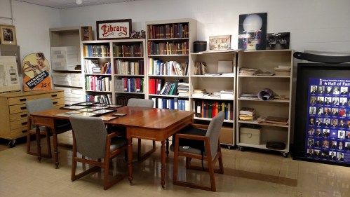 A library room with bookshelves, a wooden table, chairs, and framed photos on the walls.