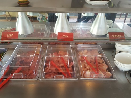 Display of raw meats labeled beef, pork, and chicken in a restaurant setting, with red tongs for serving.