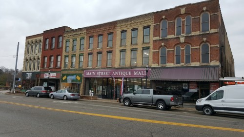 Historic brick buildings line a street, featuring shops including "Main Street Antique Mall" and parked cars.