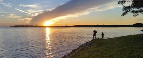 Two people fishing by a river at sunset, with vibrant colors reflecting on the water and a serene landscape.