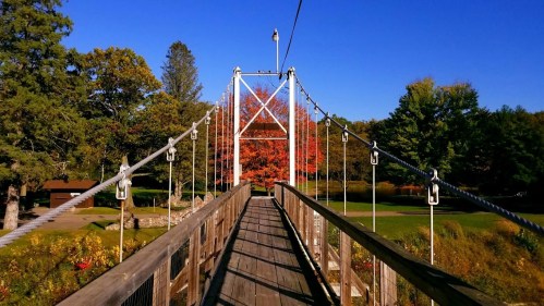A wooden suspension bridge stretches over a scenic landscape with colorful autumn trees under a clear blue sky.