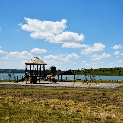 A playground with a wooden structure and swings, set against a blue sky with clouds and a lake in the background.