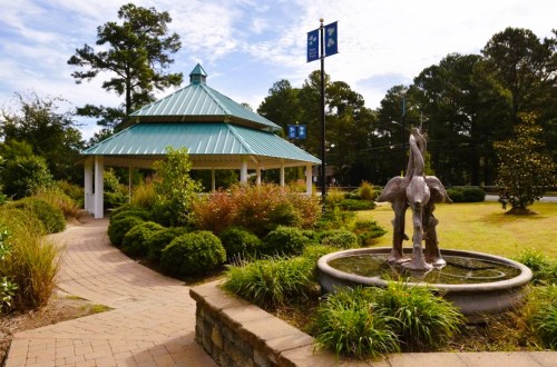 A gazebo with a green roof surrounded by lush greenery and a fountain featuring a sculpture in a park setting.