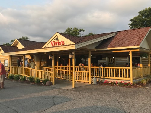 A cozy restaurant named Vivio's with a yellow exterior and a porch, surrounded by greenery and patrons enjoying the evening.