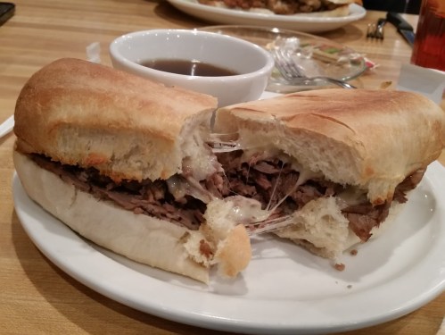 A sliced sandwich filled with beef and melted cheese, served with a small bowl of dipping sauce on a plate.