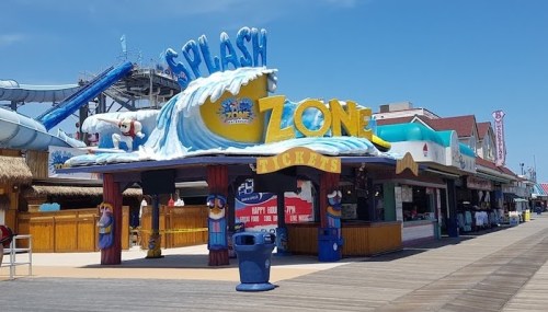 Colorful entrance to the Splash Zone water park, featuring a wave sign and ticket booth on a sunny boardwalk.