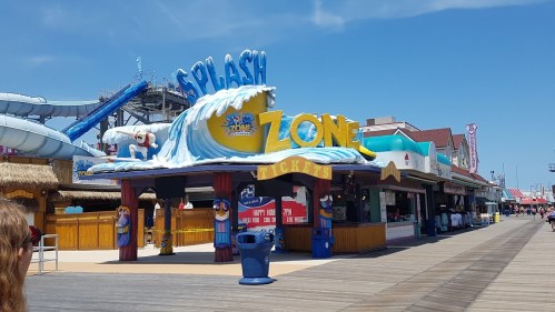 Colorful "Splash Zone" entrance on a boardwalk, with water slides in the background and clear blue skies above.