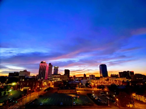City skyline at sunset, featuring colorful clouds and illuminated buildings against a deep blue sky.