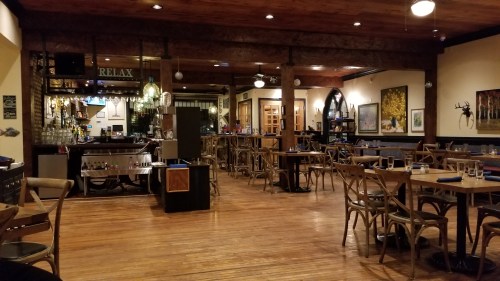 Cozy restaurant interior with wooden floors, tables set for dining, and a bar area in the background. Warm lighting.