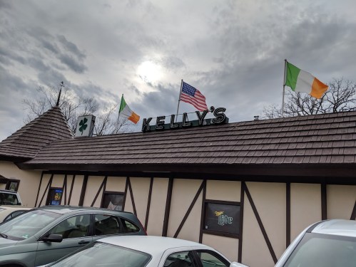 Exterior of Kelly's pub with Irish and American flags, cloudy sky, and parked cars in front.