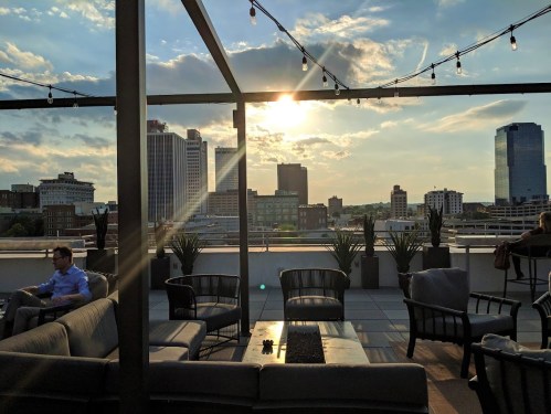A rooftop lounge with seating, overlooking a city skyline at sunset, with string lights and clouds in the sky.