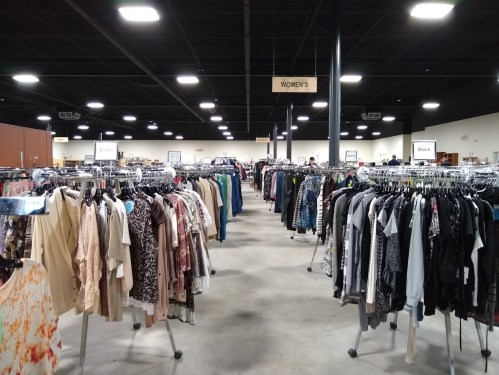 A spacious thrift store aisle filled with racks of women's clothing, organized by color and style.