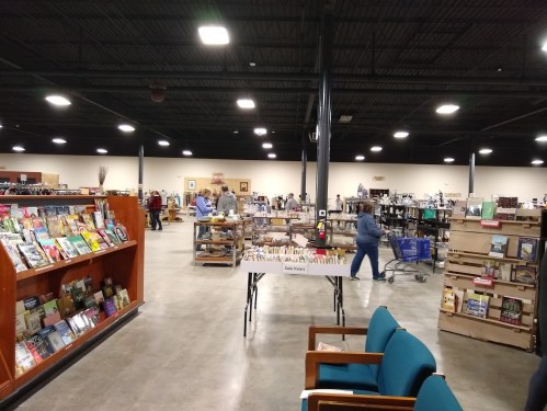 A spacious bookstore with shelves of books, shoppers browsing, and a table displaying more books in a well-lit area.