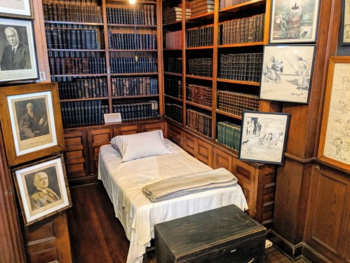A cozy library corner with a bed, bookshelves filled with books, and framed pictures on the walls.