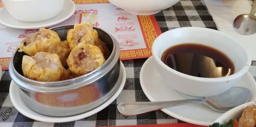 A metal steamer filled with dumplings next to a small bowl of dark sauce on a checkered tablecloth.