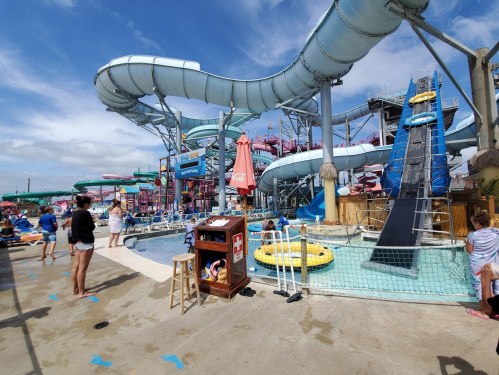 A vibrant water park scene with slides, a pool, and people enjoying the attractions under a blue sky.