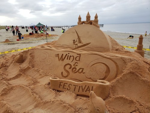Sand sculpture featuring "Wind & Sea Festival" with waves and a windsurfer, set on a beach with cloudy skies.