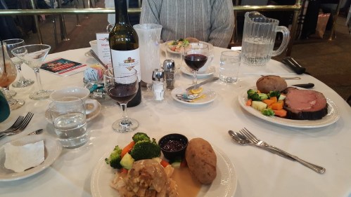A dining table set with plates of food, wine, and water glasses, featuring roast beef, vegetables, and baked potatoes.