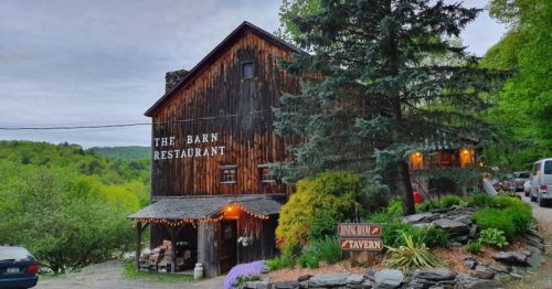 A rustic wooden barn restaurant surrounded by greenery, with a sign for a tavern and outdoor seating.