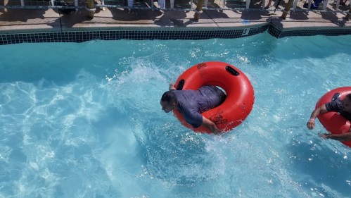 Two people in red inner tubes splash through a blue pool, enjoying a sunny day.