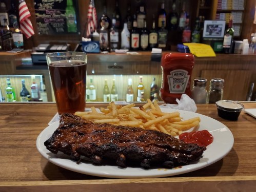 A plate of BBQ ribs with fries and ketchup, alongside a glass of dark beer, set on a wooden bar counter.