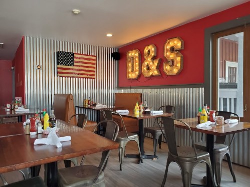 Cozy restaurant interior with metal accents, wooden tables, and an American flag on the wall, featuring bright "D&S" signage.