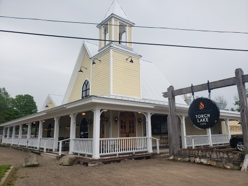 A yellow building with a steeple, featuring a porch and a sign that reads "Torch Lake Cafe."