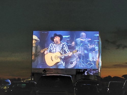 A musician performs on a large screen at dusk, with cars parked in the foreground and a band visible behind him.