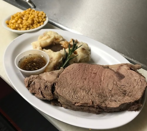 A plate with sliced roast beef, mashed potatoes with gravy, corn, and a small cup of sauce, garnished with rosemary.