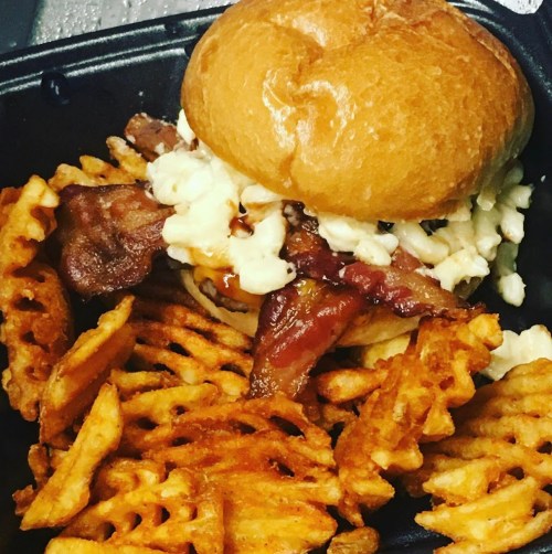 A close-up of a sandwich topped with bacon and sauce, served with a side of waffle fries in a black container.