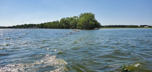 A serene lake scene with gentle waves, green trees on an island, and a clear blue sky.
