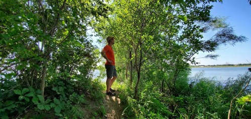 A person in an orange shirt stands by a river, surrounded by lush greenery and trees under a clear blue sky.