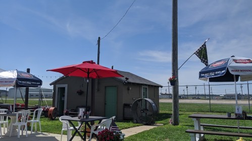 A small outdoor dining area with umbrellas, tables, and a shed, set against a clear blue sky and grassy field.