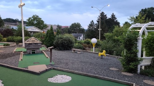 A mini-golf course with green turf, a small house structure, and a yellow dog statue amidst trees and cloudy skies.