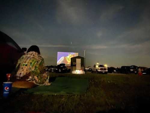 A person sits on a blanket watching a movie on a large screen outdoors at night, surrounded by parked cars.